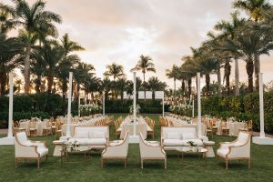 Luxury garden wedding setup with white and gold furniture, lush floral arrangements, and tropical palm tree backdrop.
