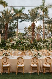 Luxury garden wedding setup with white and gold furniture, lush floral arrangements, and tropical palm tree backdrop.