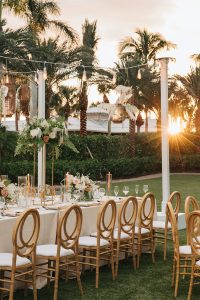 Luxury garden wedding setup with white and gold furniture, lush floral arrangements, and tropical palm tree backdrop.