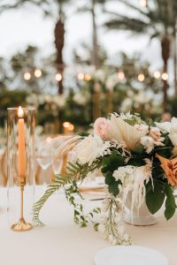 Luxury garden wedding setup with white and gold furniture, lush floral arrangements, and tropical palm tree backdrop.