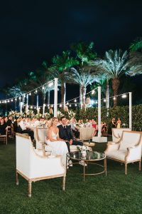 Luxury garden wedding setup with white and gold furniture, lush floral arrangements, and tropical palm tree backdrop.